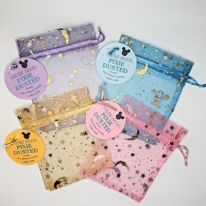May include: Five sheer organza drawstring pouches in purple, blue, gold, and pink, each featuring gold foil moon and star patterns. Attached to each pouch is a round card with a Mickey Mouse icon and the text You've Been Pixie Dusted.