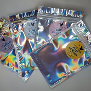 May include: Four rectangular, holographic, resealable pouches with a metallic, iridescent finish. Each pouch features a circular sticker with a mouse ear silhouette and the text: You've Been Pixie Dusted, From: The Parker Family, Cabin 5520.