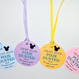 May include: Four circular paper tags in light blue, lavender, yellow, and pink, each with a matching satin ribbon. The text reads YOU'VE BEEN PIXIE DUSTED, From: The Wilson Family, Cabin 2106, accompanied by a black mouse ear silhouette icon.