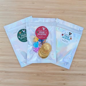 May include: Three iridescent, resealable pouches containing small items. The center pouch features a red circular sticker with the text "YOU'VE BEEN PIXIE DUSTED" and contains gold-colored coins and colorful, star-shaped confetti pieces.