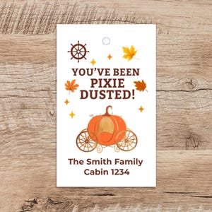 May include: A white tag featuring the text "YOU'VE BEEN PIXIE DUSTED!" and a pumpkin carriage illustration. The tag also includes the text "The Smith Family Cabin 1234". Decorative elements include a ship's wheel, leaves, and stars.
