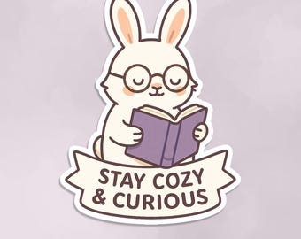 May include: A white cartoon bunny wearing glasses and reading a purple book. A banner below the bunny reads "STAY COZY & CURIOUS." The sticker has a white border and the text "ROWYAN DESIGNS" at the bottom.