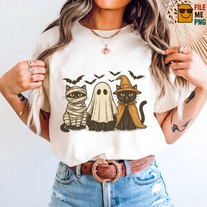 May include: Cream-colored t-shirt featuring a Halloween-themed graphic. The design includes three cats dressed as a mummy, a ghost, and a witch, with bats flying above. The graphic is in shades of black, tan, and orange.