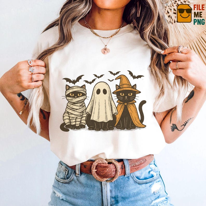 May include: Cream-colored t-shirt featuring a Halloween-themed graphic. The design includes three cats dressed as a mummy, a ghost, and a witch, with bats flying above. The graphic is in shades of black, tan, and orange.