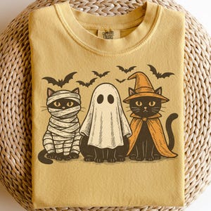 May include: Mustard-colored t-shirt featuring a Halloween design. The graphic includes a mummy cat, a ghost, and a witch cat, with bats flying above. The shirt has the words "Comfort Colors" printed on the collar.