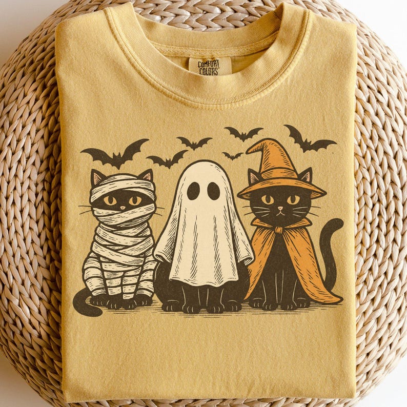 May include: Mustard-colored t-shirt featuring a Halloween design. The graphic includes a mummy cat, a ghost, and a witch cat, with bats flying above. The shirt has the words "Comfort Colors" printed on the collar.