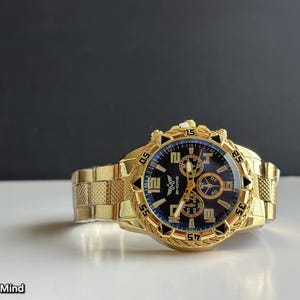 May include: A gold-toned wristwatch with a black and blue face. The watch features a textured gold band and a detailed bezel. The watch face displays multiple subdials and gold-colored hands and markers. The text "ZHONE" is visible on the watch face. The text "Choice of Mind" is visible at the bottom.