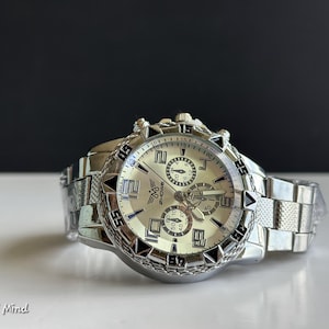 May include: Silver-toned wristwatch with a light-colored face and multiple sub-dials. The watch features a textured bezel and a linked metal band. The text "ZHOWE" is visible on the watch face. The watch is on a white surface.