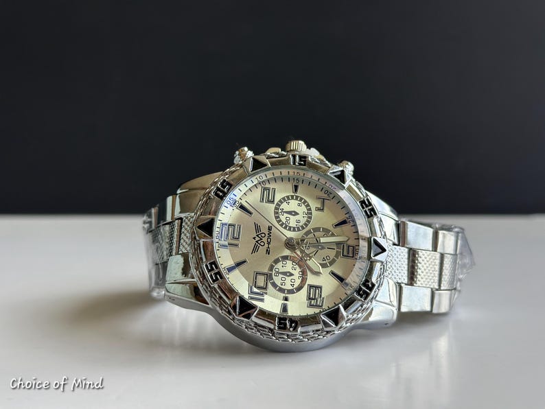 May include: Silver-toned wristwatch with a light-colored face and multiple sub-dials. The watch features a textured bezel and a linked metal band. The text "ZHOWE" is visible on the watch face. The watch is on a white surface.