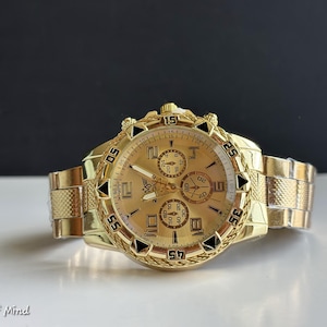 May include: A gold-toned wristwatch with a textured band and face. The watch features multiple subdials and black accents. The watch face displays the numbers 5, 15, 25, 35, 45, and 55. The watch is on a white surface with a black background.