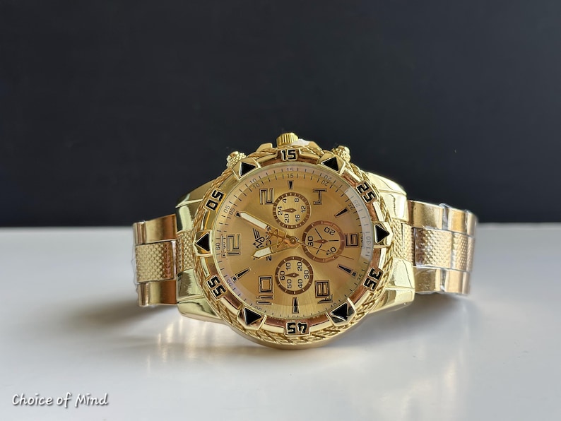 May include: A gold-toned wristwatch with a textured band and face. The watch features multiple subdials and black accents. The watch face displays the numbers 5, 15, 25, 35, 45, and 55. The watch is on a white surface with a black background.