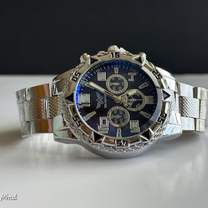 May include: A silver-toned wristwatch with a dark blue face and multiple sub-dials. The watch features a textured bezel and a stainless steel band. The watch is displayed on a white surface.
