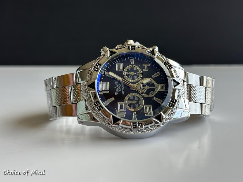 May include: A silver-toned wristwatch with a dark blue face and multiple sub-dials. The watch features a textured bezel and a stainless steel band. The watch is displayed on a white surface.