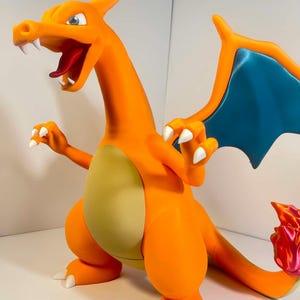 May include: A vibrant orange Charizard figurine with blue wings and white claws. The dragon-like figure has an open mouth revealing a red tongue and white teeth. The belly is a light yellow color, and the tail has a flame design.