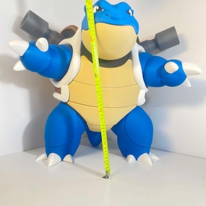 May include: A large, blue and cream-colored Blastoise figure, a popular character from the Pokémon franchise. The figure has a yellow shell, white accents, and two gray cannons on its back. A measuring tape shows the figure is approximately 20 inches tall.