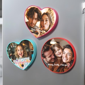 Könnte beinhalten: Drei farbige Fotorahmen mit Familienfotos werden auf einem grauen Kühlschrank angezeigt. Einer ist herzförmig mit rosa Rand, ein anderer herzförmig mit blauem Rand, und der dritte ist rund mit rotem Rand. Text ist auf den Rahmen sichtbar.