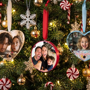 Op de afbeelding: Drie gepersonaliseerde foto-kersthangers in een kerstboom. Twee zijn hartvormig met witte en lichtblauwe randen, en één is rond met een rode rand. Elke hanger bevat een persoonlijke foto en is opgehangen aan een gekleurd lint.