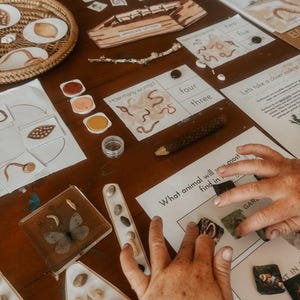 May include: A wooden table displays educational materials. There are printed cards with numbers and worms, a tray with seeds, and a butterfly encased in resin. The text "UPPERCASE LETTERS" is visible on a wooden piece.