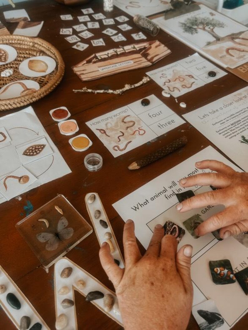 May include: A wooden table displays educational materials. There are printed cards with numbers and worms, a tray with seeds, and a butterfly encased in resin. The text "UPPERCASE LETTERS" is visible on a wooden piece.