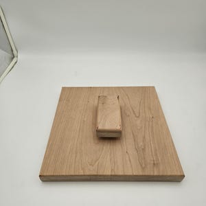 May include: A light-colored wooden cutting board with a rectangular base and a raised handle. The wood grain is visible, and the board appears to be new. The handle is a small, rectangular block.