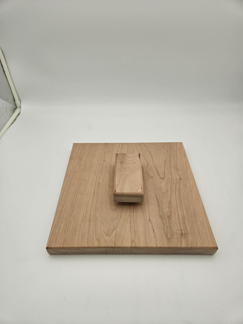 May include: A light-colored wooden cutting board with a rectangular base and a raised handle. The wood grain is visible, and the board appears to be new. The handle is a small, rectangular block.