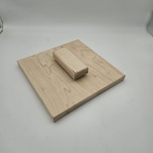 May include: A light-colored wooden square with a rectangular block on top. The wood grain is visible. The block is centered on the square. The objects are made of natural wood.