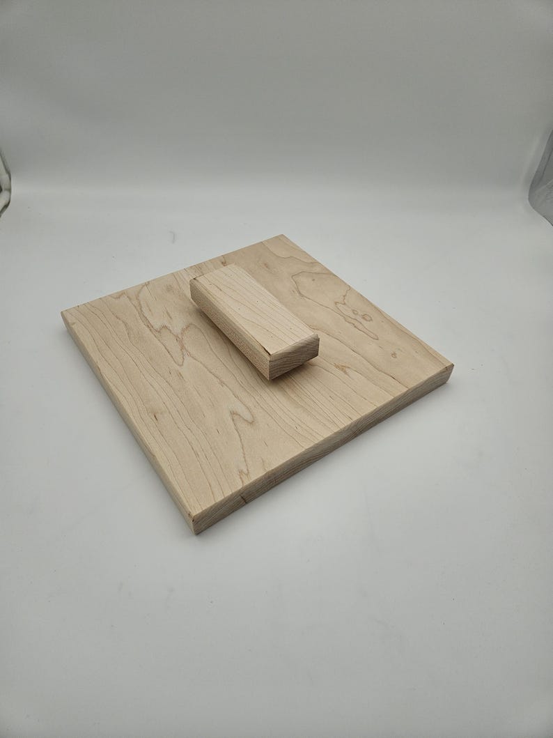 May include: A light-colored wooden square with a rectangular block on top. The wood grain is visible. The block is centered on the square. The objects are made of natural wood.