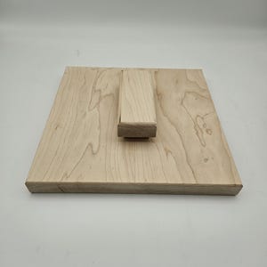 May include: A square, light-colored wooden display stand with a rectangular support. The wood grain is visible on the surface. The stand is designed to hold an item for display.