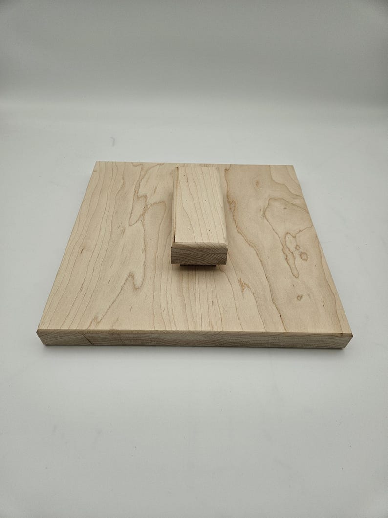 May include: A square, light-colored wooden display stand with a rectangular support. The wood grain is visible on the surface. The stand is designed to hold an item for display.