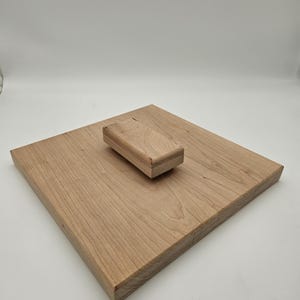 May include: A light-colored wooden cutting board with a matching rectangular wooden block. The cutting board is square-shaped and has visible wood grain. The block is smaller and rests on the board. The items are made of natural wood.