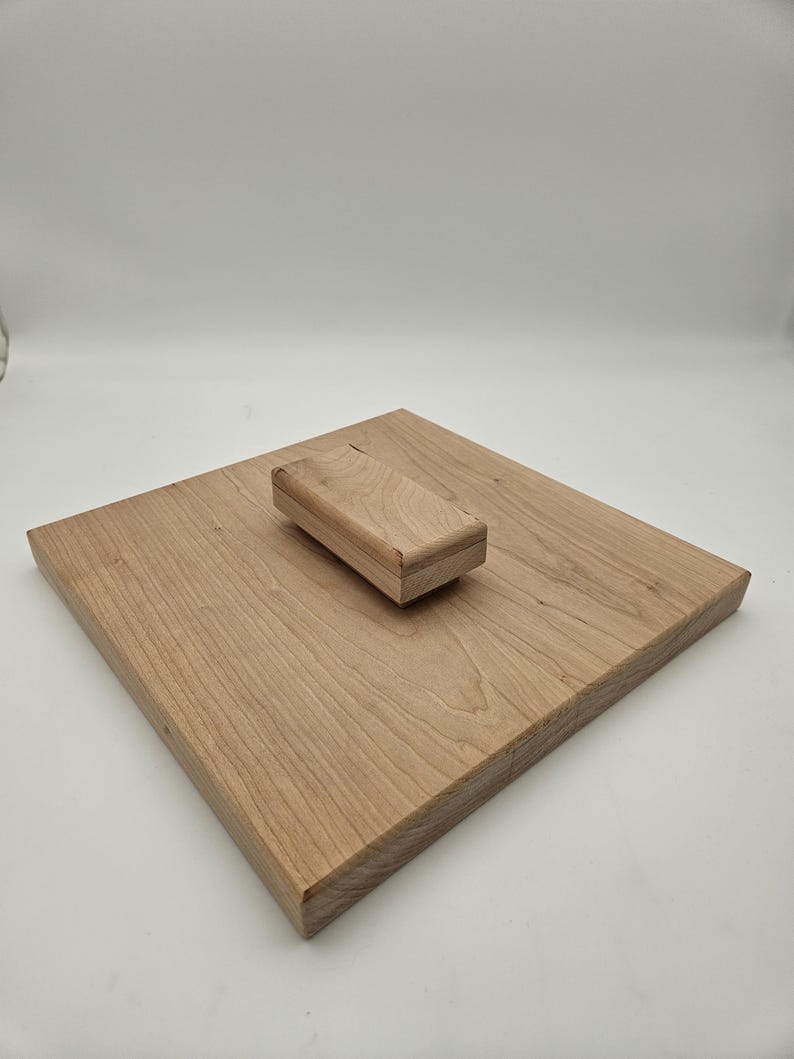 May include: A light-colored wooden cutting board with a matching rectangular wooden block. The cutting board is square-shaped and has visible wood grain. The block is smaller and rests on the board. The items are made of natural wood.