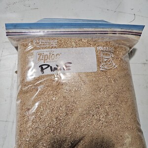 Puede incluir: Una bolsa Ziploc transparente llena de virutas de madera de color marr&oacute;n claro. La bolsa est&aacute; sellada con una cremallera azul y tiene una etiqueta blanca con la palabra "Pine".