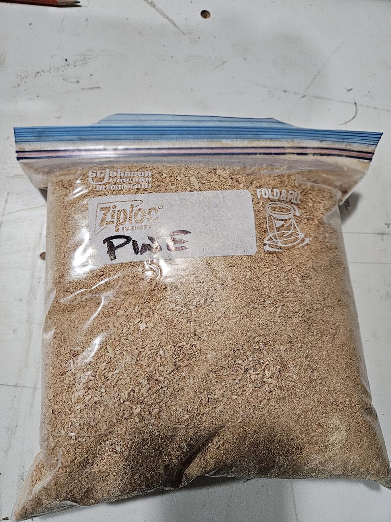 Puede incluir: Una bolsa Ziploc transparente llena de virutas de madera de color marr&oacute;n claro. La bolsa est&aacute; sellada con una cremallera azul y tiene una etiqueta blanca con la palabra "Pine".