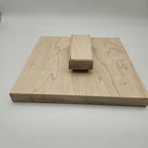 May include: A light-colored wooden cutting board with a rectangular base and a raised rectangular block. The wood grain is visible. The board is likely for kitchen use.