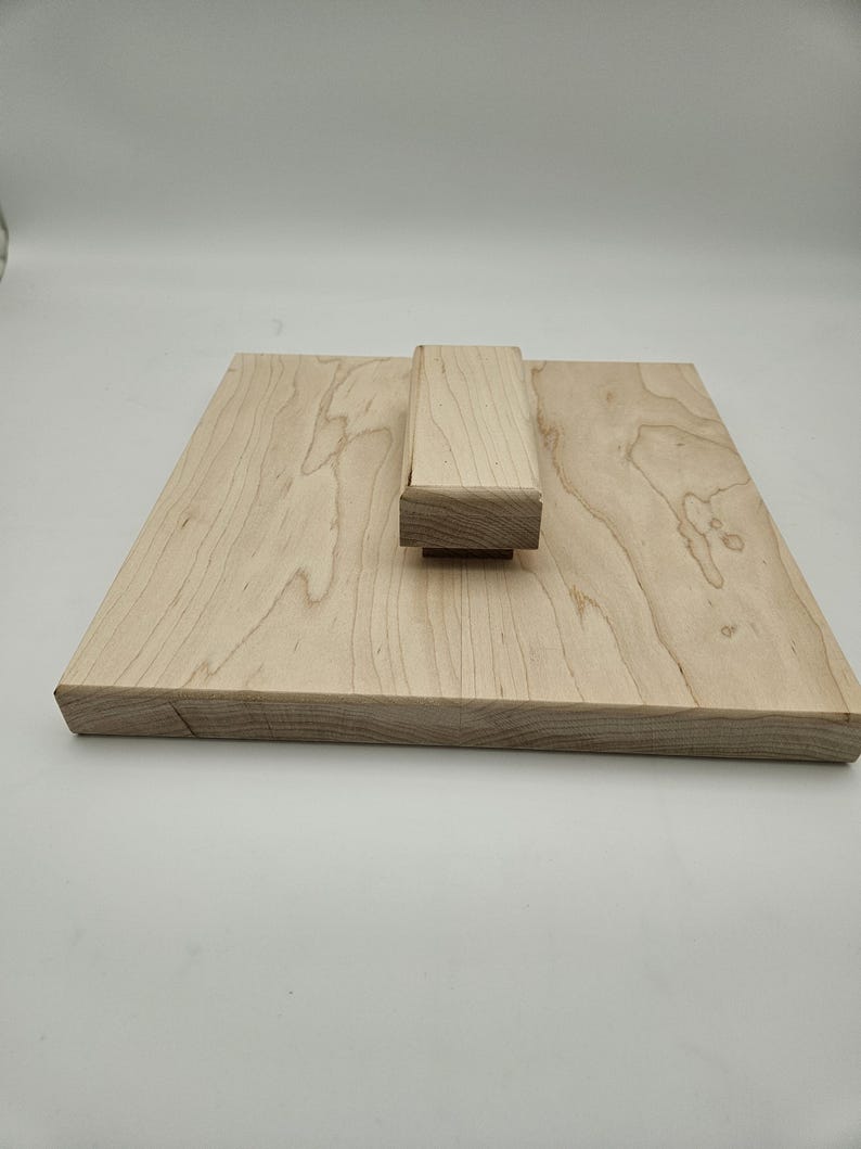 May include: A light-colored wooden cutting board with a rectangular base and a raised rectangular block. The wood grain is visible. The board is likely for kitchen use.