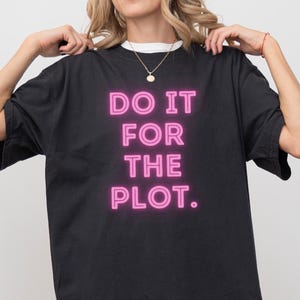 Puede incluir: Camiseta negra con la frase "DO IT FOR THE PLOT" en letras de neón rosa brillante. La camiseta tiene un corte holgado y mangas cortas. La palabra "BLACK" está impresa en la parte inferior de la camiseta.