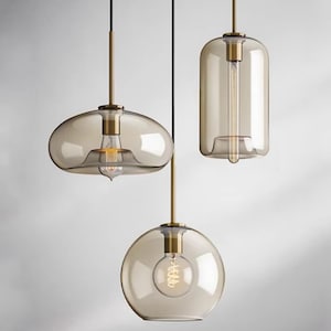 Op de afbeelding: Drie hanglampen met rookkleurige glazen kappen in verschillende vormen, waaronder een brede afgeplatte koepel, een hoge cilinder en een bol. Elk armatuur heeft een messingkleurige metalen fitting en een zichtbare filamentlamp aan een snoer.