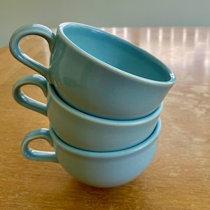 May include: A stack of three light blue ceramic cups with rounded bowls and curved handles. Each cup features a smooth, glossy finish and a consistent solid color throughout, with the top cup angled to reveal the interior basin and rim.