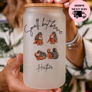 May include: A frosted glass tumbler with a bamboo lid and straw. The tumbler features a design with the text "Small, but brave" and the name "Heather". The design includes illustrations of monkeys.