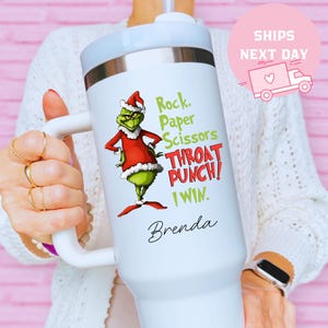 May include: White insulated tumbler with a handle, featuring a Grinch character in a Santa hat and red coat. Text reads "Rock, Paper, Scissors, Throat Punch! I Win." and "Brenda". The tumbler has a light blue lid and a silver rim.