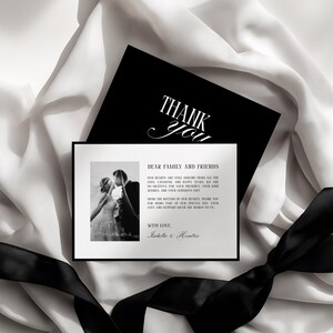 May include: A black and white wedding thank you card with a photo of a couple kissing. The card has the words "THANK YOU" in elegant script and a message to family and friends. A black ribbon and white fabric are in the background.