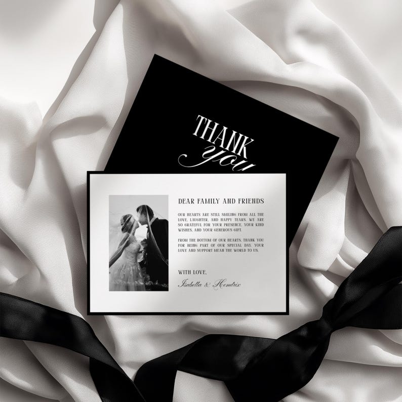 May include: A black and white wedding thank you card with a photo of a couple kissing. The card has the words "THANK YOU" in elegant script and a message to family and friends. A black ribbon and white fabric are in the background.