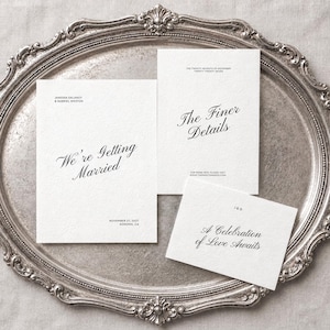 May include: Three white rectangular wedding stationery cards with black serif and script typography. The cards feature the text We're Getting Married, The Finer Details, and A Celebration of Love Awaits, along with names, dates, and a website URL.