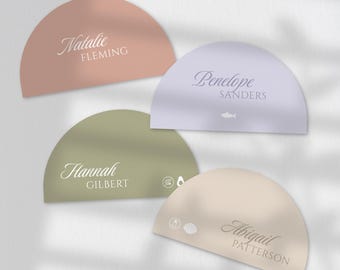 Könnte beinhalten: Vier halbkreisförmige Tischkarten in Rosa-, Lavendel-, Grün- und Beigetönen. Jede Karte trägt einen Vor- und Nachnamen in eleganter Schrift. Die Karten haben ein minimalistisches Design, mit kleinen Symbolen auf zwei der Karten.