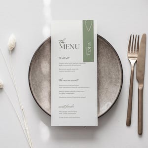 Op de afbeelding: Een menukaart met de tekst "the MENU" en een lijst met gerechten, waaronder Caprese salade en kipfilet met kruiden. De kaart ligt op een grijze bord met een vork en mes. Het menu heeft een groene accent met de naam "ALICIA".
