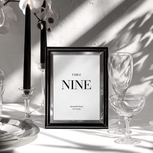 以下が含まれることがあります： ミニマリストフォントで「NINE」という文字が入った黒いフレームのテーブルナンバーサイン。サインは白い背景で、「Kendall & Ryan」という名前と「09/19/2028」という日付が含まれています。テーブルセッティングにはガラス製品と黒いキャンドルが含まれています。