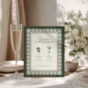 May include: A dark green wedding cocktail menu with a white lace border. The menu features two cocktail illustrations and the words "Signature Cocktails". A champagne flute is in the foreground.