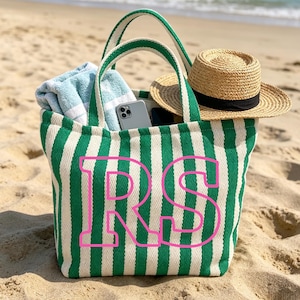 May include: A green and white striped tote bag with the pink letters "RS" on the front. The bag contains a blue towel, a smartphone, and a straw hat with a black band. The bag is on a sandy beach.