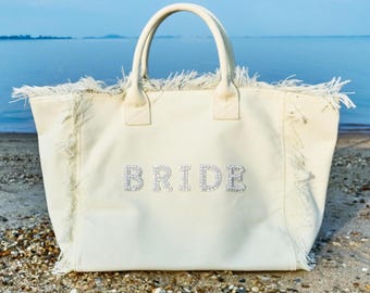 May include: Cream-colored tote bag with the word "BRIDE" spelled out in sparkling rhinestones. The bag has two handles and a fringed edge. The background shows a beach and ocean.