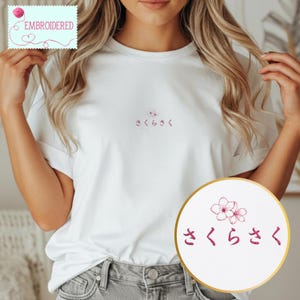 May include: White t-shirt with embroidered cherry blossom design and Japanese text. The design includes a small flower and the Japanese characters "さくらさく" in a pink color. The shirt has a classic crew neck and short sleeves.